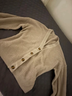 Beige stickad kofta med knappar - Säljer en beige stickad kofta med snygga bruna knappar framtill. Koftan har långärmad design och är ribbstickad för en chill och mysig känsla. Perfekt att slänga på sig när det är lite kyligt ute. Färgen på koftan är jätte 🤩. Den formar överkroppen så de ser perfekt ut. Inget är fel på plagget utan jag rensar lite😊