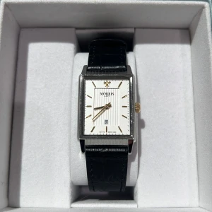 Svart läderklocka från Morris - Stilren klocka från Morris med rektangulär urtavla i vitt och gulddetaljer. Svart läderarmband och spänne i silver. Helt oanvänd, prislapp kvar (ordinarie pris 1799;-) 