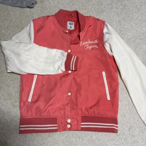 Orange och vit bomberjacka Crocker S - Säljer en snygg orange och vit bomberjacka från Crocker i storlek S. Jackan har vita ärmar, broderad text på bröstet och klassiska ränder på muddar och krage. Stängs med tryckknappar och har två fickor fram (obs: ej riktiga fickor, endast för design). Perfekt för dig som vill ha en retro college-look.