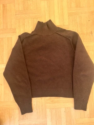 Brun polotröja i ull - Mysig brun polotröja med ribbad hög krage och muddar. Tröjan är stickad i ull och har en loose passform med långa ärmar. Perfekt för kyliga dagar när du vill vara både varm och stilren.