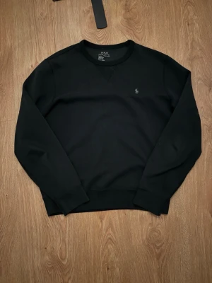Svart sweatshirt från Polo Ralph Lauren - En stilren svart sweatshirt från Polo Ralph Lauren med klassisk rund hals och broderad logga på bröstet. Tröjan har långa ärmar och är gjord i mjuk bomull, perfekt för en clean och avslappnad look.