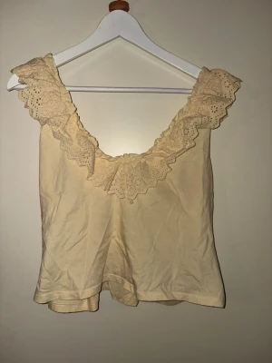 Gul linnetopp med spets från Gina Tricot - Supersöt beige linnetopp från Gina Tricot med breda axelband och vacker spetsvolang runt urringningen. Toppen är ärmlös och har en mjuk, lätt A-linjeformad passform. Perfekt för varma dagar och enkel att styla.