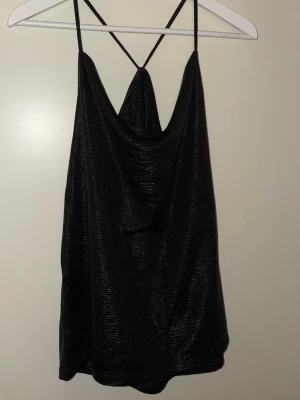 Fest topp - Säljer en svart glittrig linnetopp från Soaked in Luxury. Toppen har tunna axelband som korsas i ryggen och en snygg draperad front. Materialet är glansigt och ger en lyxig känsla, perfekt för att sticka ut. Passar dig som gillar trendiga detaljer och vill ha något extra.