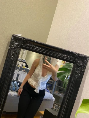 Vit spetsig omlottopp med volang - Säljer en superfin vit omlottopp i spets med volangdetaljer och djup v-ringning. Toppen är ärmlös, har öppen rygg och knyts med långa band bak. Sitter tight och croppad, perfekt till byxor eller kjol. 