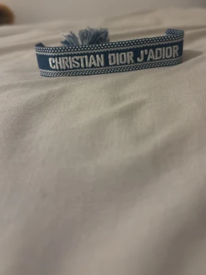 Blått Christian Dior J'Adior armband - Säljer ett blått tygarmband från Christian Dior med texten 'J'Adior' broderat i vitt. Armbandet har justerbar knytning med tofsar i ändarna och en snygg, ungdomlig design som passar perfekt till din outfit.pris kan diskuteras 
