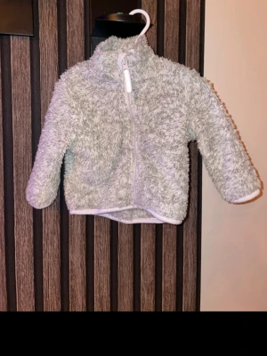 Ljusgrå teddyjacka med dragkedja storlek 68 - Supermjuk och fluffig teddyjacka i ljusgrått med hel dragkedja framtill och hög krage. Perfekt för kyliga dagar och riktigt mysig att ha på sig. Jackan har långa ärmar och en enkel, stilren look. Storlek 68