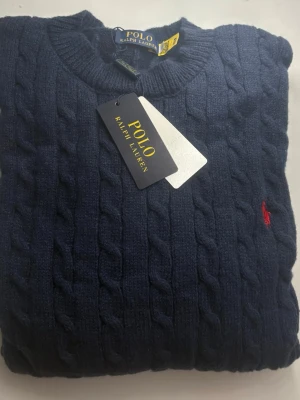 Mörkblå kabelstickad tröja - Kabelstickad mörkblå tröja med klassisk rund halsringning och den ikoniska röda logon broderad på bröstet. Tröjan har långa ärmar och ribbade muddar. Perfekt för dig som vill ha en stilren och tidlös look.