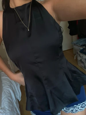 Svart satinlinnetopp med utsvängd midja - Säljer en svart linnetopp i satin med hög hals och ärmlös design. Toppen har en snygg utsvängd midja som ger en feminin siluett och en elegant look. Perfekt att styla med jeans eller kjol för en trendig outfit.