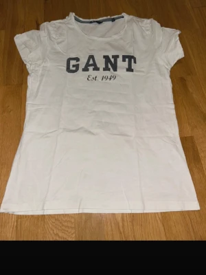 Vit GANT t-shirt med tryck - Säljer en klassisk vit t-shirt från GANT i storlek M. T-shirten har korta ärmar och ett stort mörkblått GANT-tryck med 'Est. 1949' på bröstet. Tillverkad i mjuk bomull och har en rund halsringning med randig detalj på insidan.