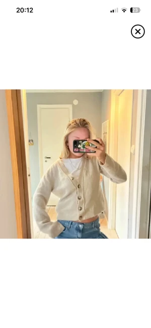 Beige kofta med knappar och v-ringning - Mysig beige kofta med v-ringning och stora, mönstrade knappar framtill. Koftan är lite croppad och har långa ärmar. Perfekt att slänga över en t-shirt och matcha med jeans för en chill look. Storlek s men passar även xs för den e liten.