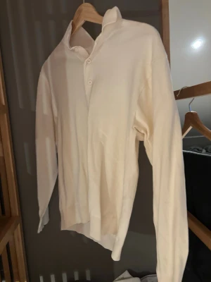 Vit polotröja med knappar - Klassisk vit polotröja med lång ärm och tre knappar vid halsen. Tröjan har en enkel och stilren design med ribbade muddar vid ärmslut och nederkant. Perfekt för lager på lager och passar till det mesta i garderoben.