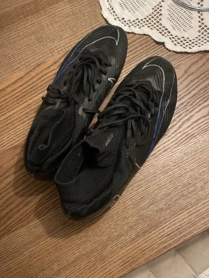 Nike Mercurial svarta fotbollsskor - Säljer ett par Nike Mercurial fotbollsskor i svart med blå och vita detaljer. Skorna har snörning, högre strumpliknande skaft och är tillverkade i syntetmaterial för bästa grepp och känsla på planen. Perfekta för dig som vill sticka ut med stil på matchen.