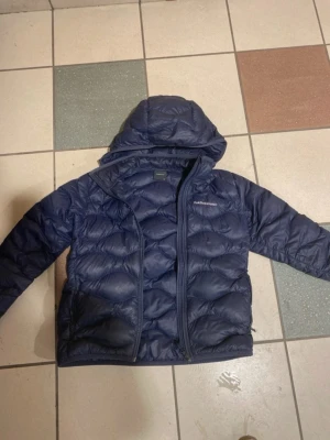 Mörkblå dunjacka från Peak Performance - Säljer en mörkblå dunjacka från Peak Performance med huva och dragkedja framtill. Jackan är quiltad och har en clean look som passar perfekt till kalla dagar. Klassisk modell med lång ärm och diskret logga på bröstet.