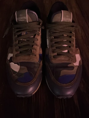 Valentino Garavani camo sneakers - Säljer ett par Valentino Garavani sneakers med camouflage-mönster i grönt, blått, beige och vitt. Skorna har snörning, rund tå och detaljer i mocka och skinn. Sulan är grovmönstrad och hälen har en cool blå detalj. Perfekta för dig som vill sticka ut med lyxig streetstyle.