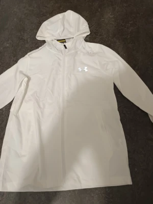 Vit vindjacka Under Armour  - Säljer en vit vindjacka Under Armour i storlek M. Jackan har huva, dragkedja framtill och reflekterande logga på bröstet samt stor logga på ryggen. Perfekt för löpning eller träning utomhus.