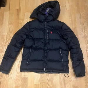 Svart pufferjacka - Svart pufferjacka med röd broderad logga på bröstet. Jackan har huva, flera dragkedjefickor och quiltad design. Materialet är syntet och dun, perfekt för kalla dagar. Stängs med dragkedja framtill och har en clean, modern look.