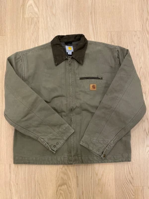 Light khaki Carhartt workwear jacka M - Snygg light khaki (beige) workwear jacka från Carhartt med brunt krage, dragkedja framtill och bröstficka med zip. Insidan har rutigt foder i mörka toner. Klassisk Carhartt-logga på bröstet och robust bomullsmaterial. Perfekt för dig som gillar utility-stil.