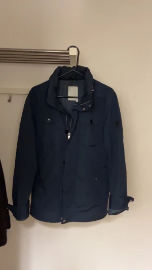 Field jacka från J.Lindeberg - Säljer min mörkblåa field jacka från J. Lindberg i ett fint skick. Jackan är i storlek M och har ett litet jack på högerarmen( kolla sista bilden) men som man inte ser när man använder den. Skriv om ni har några funderingar!