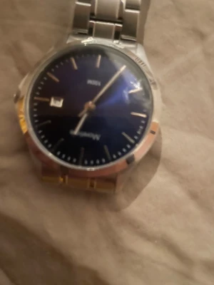 Blå och silverfärgad klocka från Moretime - Snygg klocka från Moretime med mörkblå urtavla och silverfärgat armband i metall. Urtavlan har minimalistiska markeringar och datumvisning. Länken är klassisk och robust, perfekt för dig som gillar stilrena accessoarer.