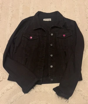 Svart jeansjacka - Svart jeansjacka med coola slitningar. Jackan har klassisk krage, knäppning framtill och fickor med rosa knappar som sticker ut. Perfekt för dig som vill ha en edgy och trendig look. Står storlek M men skulle säga S-XS