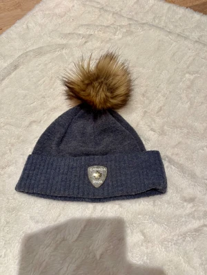 Gråblå mössa med pälsboll från Canada Snow - Mysig gråblå mössa från Canada Snow med bred ribbad kant och stor brun pälsboll på toppen. Märke med logga framtill. Perfekt för kalla dagar och snygg till vinterlooken.