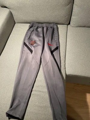 Grå PSG x Nike träningsbyxor - Snygga grå träningsbyxor från Nike med Paris Saint-Germain-logga och röd swoosh på framsidan. Byxorna har elastisk midja, svarta detaljer vid fickorna och är tillverkade i ett mjukt, sportigt material. Perfekta för dig som gillar fotboll och streetwear.