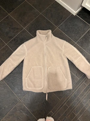 Beige teddyjacka med dragkedja - Mysig beige teddyjacka med hög krage och två stora fickor framtill. Jackan har hel dragkedja och är tillverkad i fluffigt fleece-material. Perfekt för lager-på-lager och chill dagar. Snygg loose passform och långärmad modell.