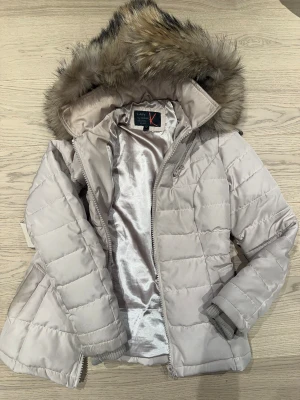 Varm jacka vinterjacka pufferjacka med äkta päls från (Lady by) Kamora  - Varm vinterjacka från Lady by Kamora med äkta päls på huvan och quiltad design. Kommer med skärp men kan användas utan. Jackan har dragkedja framtill, dold innerficka, avtagbar luva och ribbade muddar och ett glansigt innerfoder. Perfekt för kalla dagar och har en normal passform. - avtagbar luva