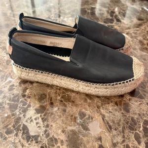 Svarta espadrillos med platåsula - Snygga svarta espadrillos med bred platåsula i flätad jute. Skorna har rund tå och ovandel i svart skinnliknande material. Perfekta för en chill sommarlook och lätta att bara dra på sig.