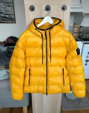 Gul pufferjacka från Replay - Fet gul pufferjacka från Replay med svart logga på ärmen och stor Replay-tryck på ryggen. Jackan har huva, svarta dragkedjor och snörning vid halsen. Insidan är röd och jackan är vadderad för extra värme. Perfekt för dig som vill sticka ut i vinter.