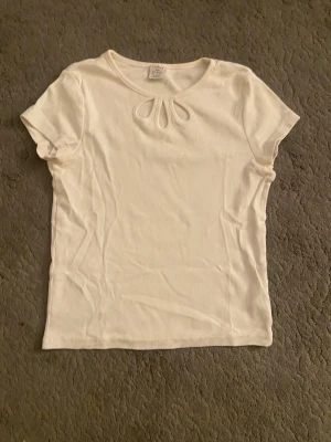 Vit ribbad topp med cut out från Lindex - Vit ribbad topp från Lindex med korta ärmar och snygga cut out-detaljer vid halsringningen. T-shirten har en enkel och fräsch look som passar perfekt till jeans eller kjol. Tillverkad i mjuk bomull för en skön känsla hela dagen.