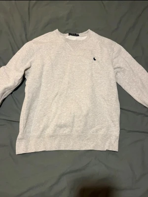 Grå sweatshirt från Polo Ralph Lauren - Snygg grå sweatshirt från Polo Ralph Lauren med klassisk rund hals och broderad marinblå logga på bröstet. Tröjan har långa ärmar och ribbade muddar vid hals, ärmslut och nederkant. Perfekt för en chill och stilren look.
