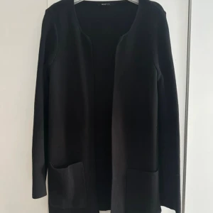 Svart cardigan jacka med fickor från Gina Tricot - Svart cardigan jacka med lång ärm och två stora fickor framtill. Enkel och stilren design utan knappar eller dragkedja, perfekt för lager-på-lager och chill vibe. Enkel och stilren design som passar till många olika outfits