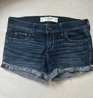 Mörkblå jeansshorts Abercrombie & Fitch - Snygga mörkblå jeansshorts från Abercrombie & Fitch med klassisk femficksdesign och fransiga kanter. Shortsen har gulbruna sömmar, knapp och dragkedja framtill samt broderade detaljer på bakfickorna. 