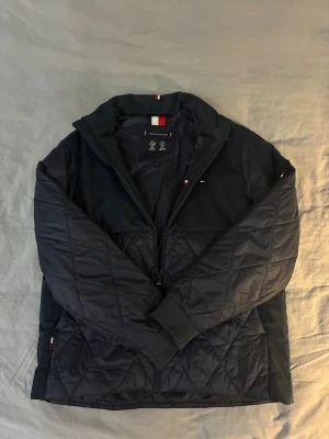 Svart quiltad jacka Tommy Hilfiger - Snygg marinblå quiltad jacka från Tommy Hilfiger med dragkedja framtill och diskreta logodetaljer. Jackan har lång ärm, ribbade muddar och klassisk krage. Materialet är polyester och designen är stilren med diamantquiltning på nedre delen och ärmarna. Nypris:1200kr Mitt pris:499kr
