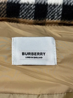 Beige pufferväst från Burberry - Snygg beige pufferväst från Burberry med ikoniskt rutigt foder i huvan. Västen har svarta dragkedjor på fickorna, tryckknappar och stor Burberry-logga på insidan. Perfekt för lager-på-lager och har en clean, modern look.