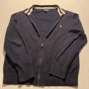 Marinblå kofta från Polo Ralph Lauren - Snygg marinblå kofta från Polo Ralph Lauren med dragkedja framtill och vit broderad logga på bröstet. Kragen har coola vita ränder och tröjan är stickad i mjukt material. Perfekt för dig som gillar klassisk stil med en sportig touch. Lite fläckar vid dragkedjan.