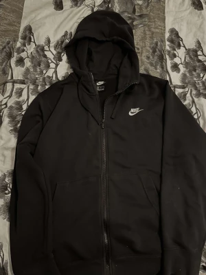 Svart Nike hoodie med dragkedja - Svart hoodie från Nike i storlek M med klassisk logga broderad på bröstet. Tröjan har huva med snörning, två fickor framtill och hel dragkedja. Perfekt för chill eller träning, mjukt bomullsmaterial och stilren look.