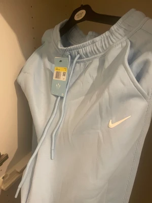 Nike NOCTA ljusblå mjukisbyxor S - Ljusblå mjukisbyxor från Nike NOCTA med vit logga på benet och Nike Swoosh på låret. Byxorna har elastisk midja med snörning, dragkedjor vid fickorna och en clean, modern look. Perfekt för chill eller streetwear.
