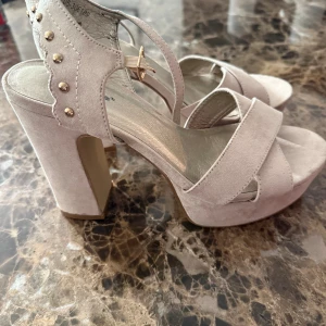 Beige sandaler med klack och nitar - Snygga beige sandaler med hög klack och plattform. Skorna har breda remmar i mocka, öppen tå och dekorativa guldfärgade nitar vid hälen. Perfekta för dig som vill sticka ut med en trendig och chic look.