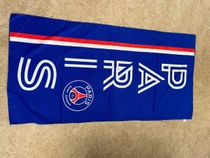 Blå PSG halsduk med logga - Snygg blå halsduk från Paris Saint-Germain med klubbens logga och vit text. Halsduken har en röd och vit rand längs ena sidan som ger en sportig vibe. Perfekt för dig som vill visa ditt stöd för PSG på matchen eller i vardagen.