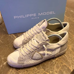 Vita Philippe Model  - Snygga vita sneakers från Philippe Model Paris med klassisk låg profil och rund tå. Skorna har detaljer i mocka, snörning och en unik logotyp på sidan. Sulan är platt och har tydlig branding undertill. Perfekt för dig som gillar stilrena och exklusiva sneakers.