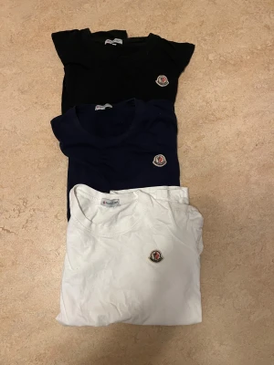 Moncler t-shirt i svart, marinblå och vit - Tre klassiska t-shirts från Moncler i svart, marinblå och vit. Alla har rund halsringning och Monclers ikoniska logga broderad på bröstet. Tillverkade i mjuk bomull för en skön och stilren look. Perfekt basplagg för garderoben.