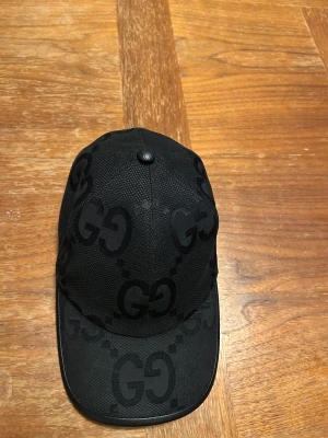 Svart Gucci keps med GG-mönster - Snygg svart keps från Gucci med det klassiska GG-mönstret över hela kepsen. Kepsen har böjd skärm och justerbar rem baktill. Materialet är slitstark polyester och designen är riktigt clean och lyxig, perfekt för dig som gillar streetwear med designer-vibe.