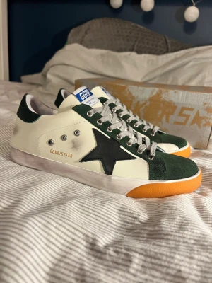 Golden Goose sneakers med grön mocka - Snygga Golden Goose sneakers i vitt skinn med gröna mockadetaljer och svart stjärna på sidan. Skorna har grå snörning, orange gummisula och GGDB/SSTAR-tryck i guld. Klassisk streetstil med slitna detaljer och låg profil. Obs: lnte 0rglnal‼️Men skorna otroligt snygga och gjorda i mycket kvalitet. Skostorlek 44 med innersulans mått på ca 28 cm. HELT NYA