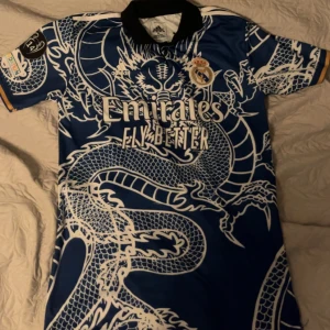 Real Madrid blå draktröja Adidas - Säljer en fet Real Madrid fotbollströja från Adidas i blått med ett stort vitt drakmönster över hela tröjan. Tröjan har svart krage, korta ärmar med gul detalj och klassiska klubbmärken samt sponsortryck på bröstet. Perfekt för dig som vill sticka ut på läktaren eller träningen.