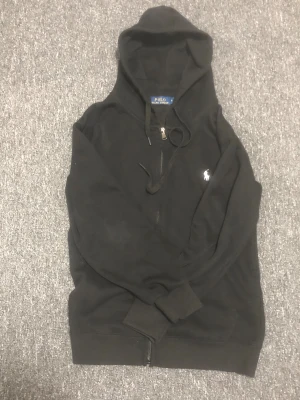 Svart hoodie från Polo Ralph Lauren - Svart hoodie från Polo Ralph Lauren i storlek S. Klassisk design med huva, snörning och dragkedja framtill. Tröjan har det ikoniska vita Polo-logot broderat på bröstet. Perfekt för chill dagar och enkel att matcha med jeans eller joggers.