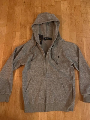 Grå hoodie från Polo Ralph Lauren - Snygg grå hoodie från Polo Ralph Lauren med klassisk logga på bröstet. Tröjan har huva med snörning, dragkedja framtill och två fickor. Tillverkad i mjuk bomullsmix och har ribbade muddar vid ärmslut och nederkant. Perfekt för chill dagar.