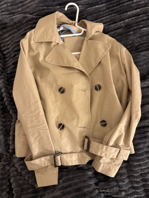 Kort beige trenchcoat med bälte - Snygg kort trenchcoat i beige med bred krage och dubbla knapprader. Jackan har bälten vid ärmsluten och midjan för extra stilpoäng. Perfekt för dig som gillar klassisk look med modern twist. Materialet känns mjukt och lätt, och knapparna har en cool marmorerad finish.