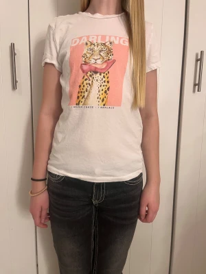 Vit t-shirt med leopardtryck - Vit t-shirt med korta ärmar och tryck framtill av en leopard med rosa bakgrund och texten 'DARLING'. T-shirten har rund hals och är gjord i mjuk bomull. Passformen är normal och trycket ger en cool och lekfull vibe, storlek 158/164